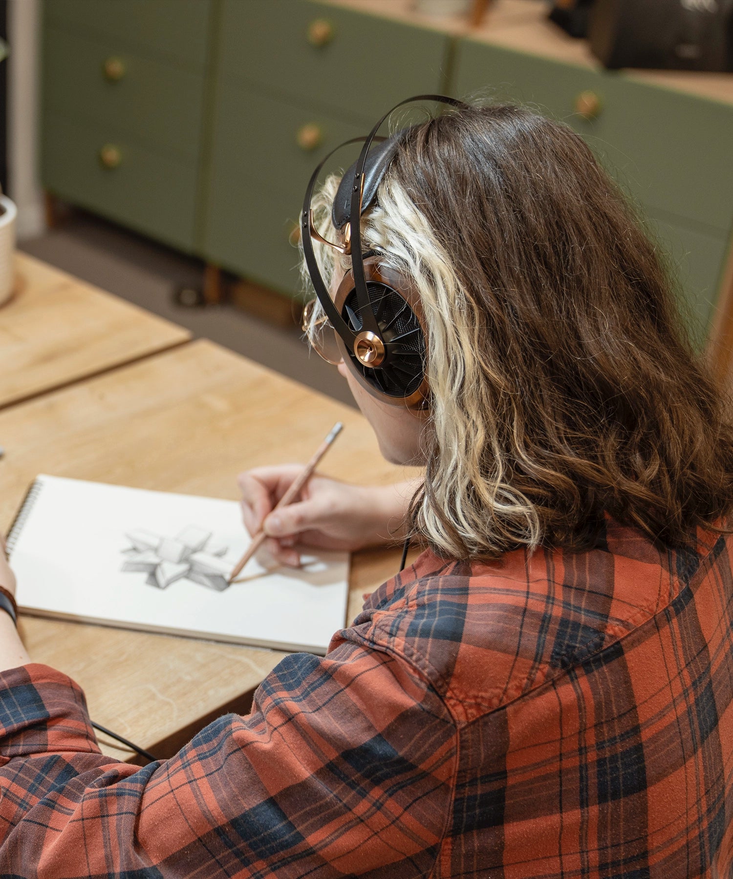 Woman in flannel shirt sitting at a desk and drawing while listening to music on her Meze Audio 109 PRO wired head phones